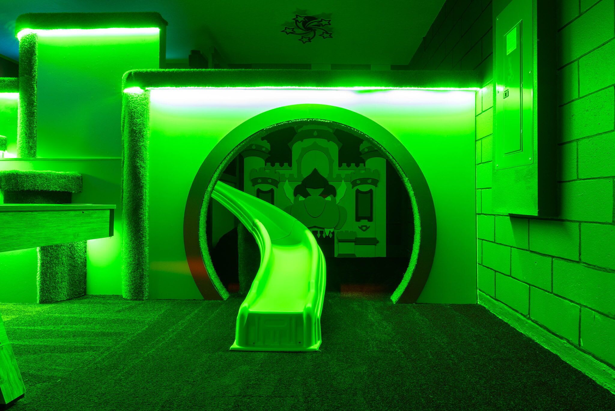 Un túnel iluminado en verde que conduce a una habitación con temática de castillo, con un tobogán, una recepción y un teléfono en la pared.