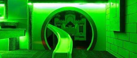 Un túnel iluminado en verde que conduce a una habitación con temática de castillo, con un tobogán, una recepción y un teléfono en la pared.
