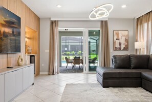 Ein modernes Wohnzimmer mit einem Ecksofa, einem Flachbildfernseher, einem Essbereich mit Stühlen und einer Glastür, die zu einer Außenpatio führt.