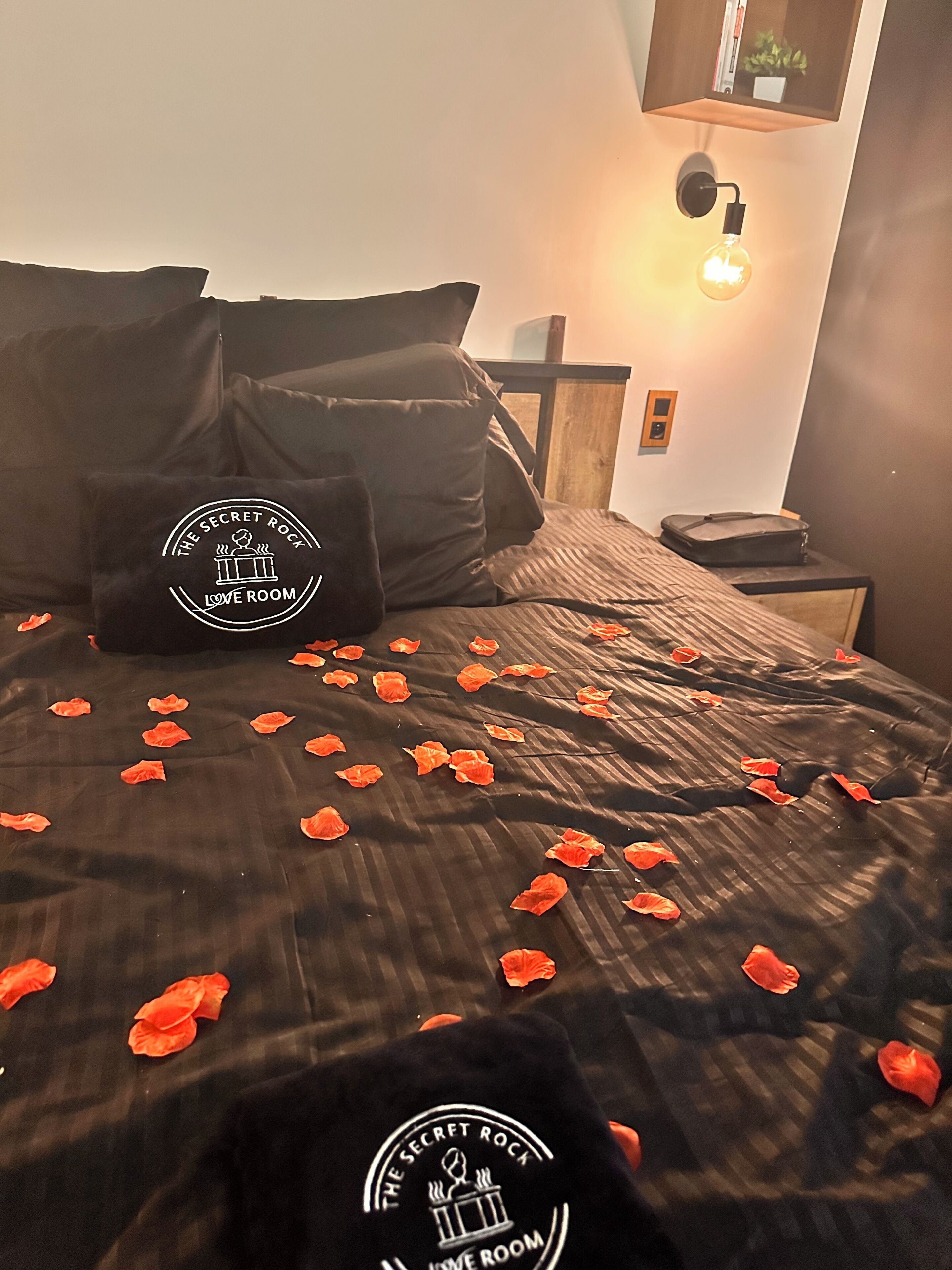 A bed with rose petals, a pillow with a logo, and a wall-mounted light fixture.