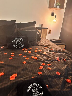 Ein Bett mit Rosenblättern, ein Kopfkissen mit einem Logo, eine Wandleuchte und ein kleiner Tisch mit einer Tasche.
