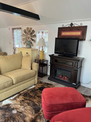A living room with a beige sofa, a red ottoman, a television, a fireplace, and a decorative wall piece. -  ROOMY SUNSHINE CABIN IN THE COUNTRY (Claremore)