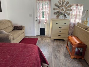 Una habitación con un sofá color beige, una colcha de cama con estampado rojo, un armario de madera y una radio antigua.
