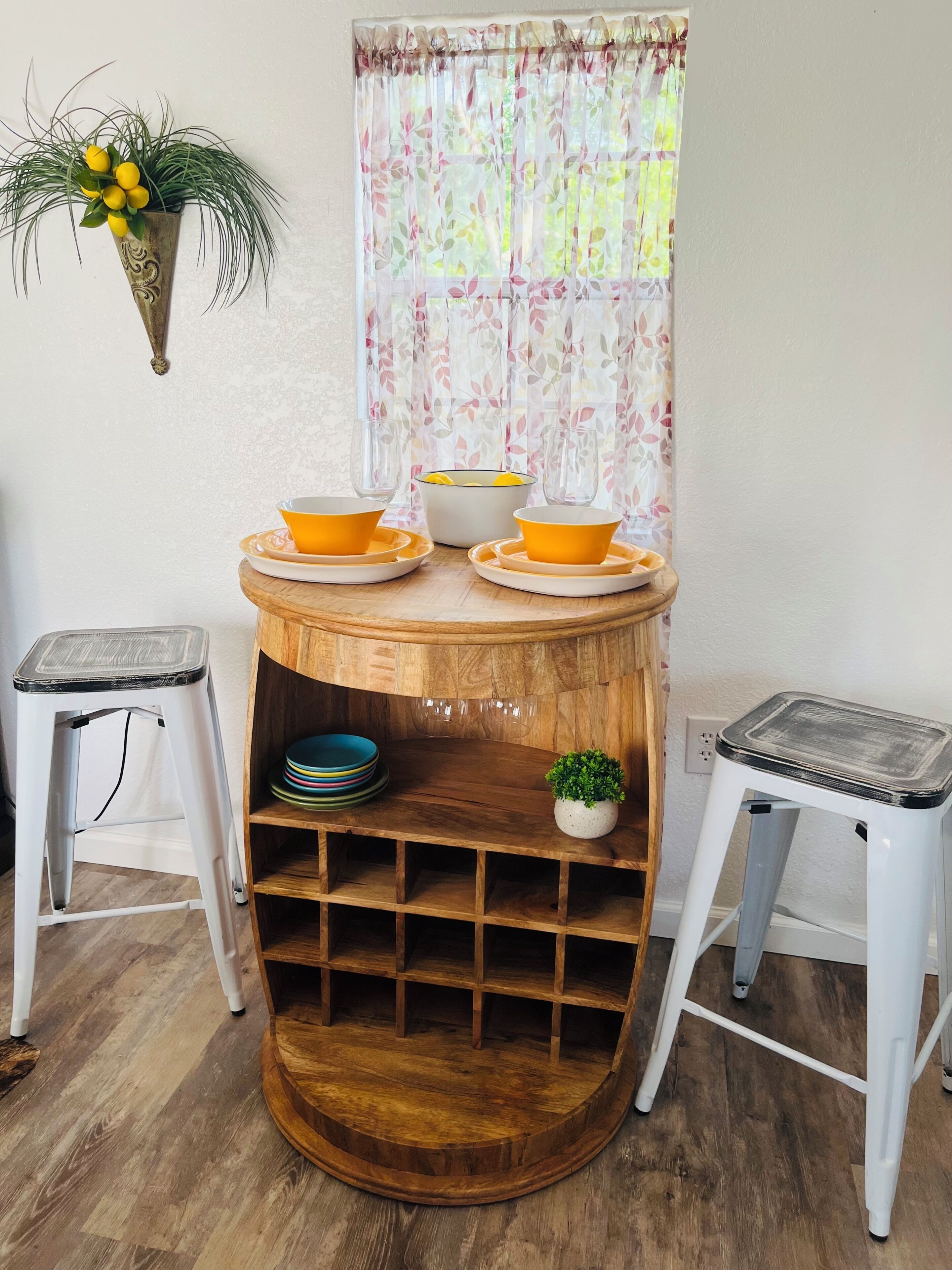 Ein Holztisch mit Ablagefläche, zwei Hocker, ein Blumentopf und ein Fenster mit blumigem Vorhang.