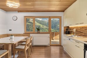 Eine moderne Küche mit Holzausstattung, ein Essbereich mit Tisch und Stühlen sowie ein Blick auf eine Berglandschaft durch große Fenster.