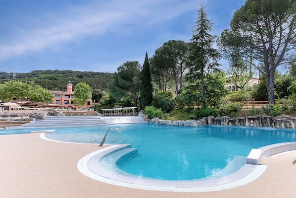 A swimming pool with a stone border, lounge chairs, and a building with a clock tower in the background. - Family Apartment for 4 in Ardeche (Grospierres)