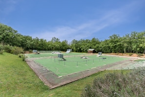A green outdoor tennis court with a fence, trees, and a small building in the background. - Family Apartment for 4 in Ardeche (Grospierres)
