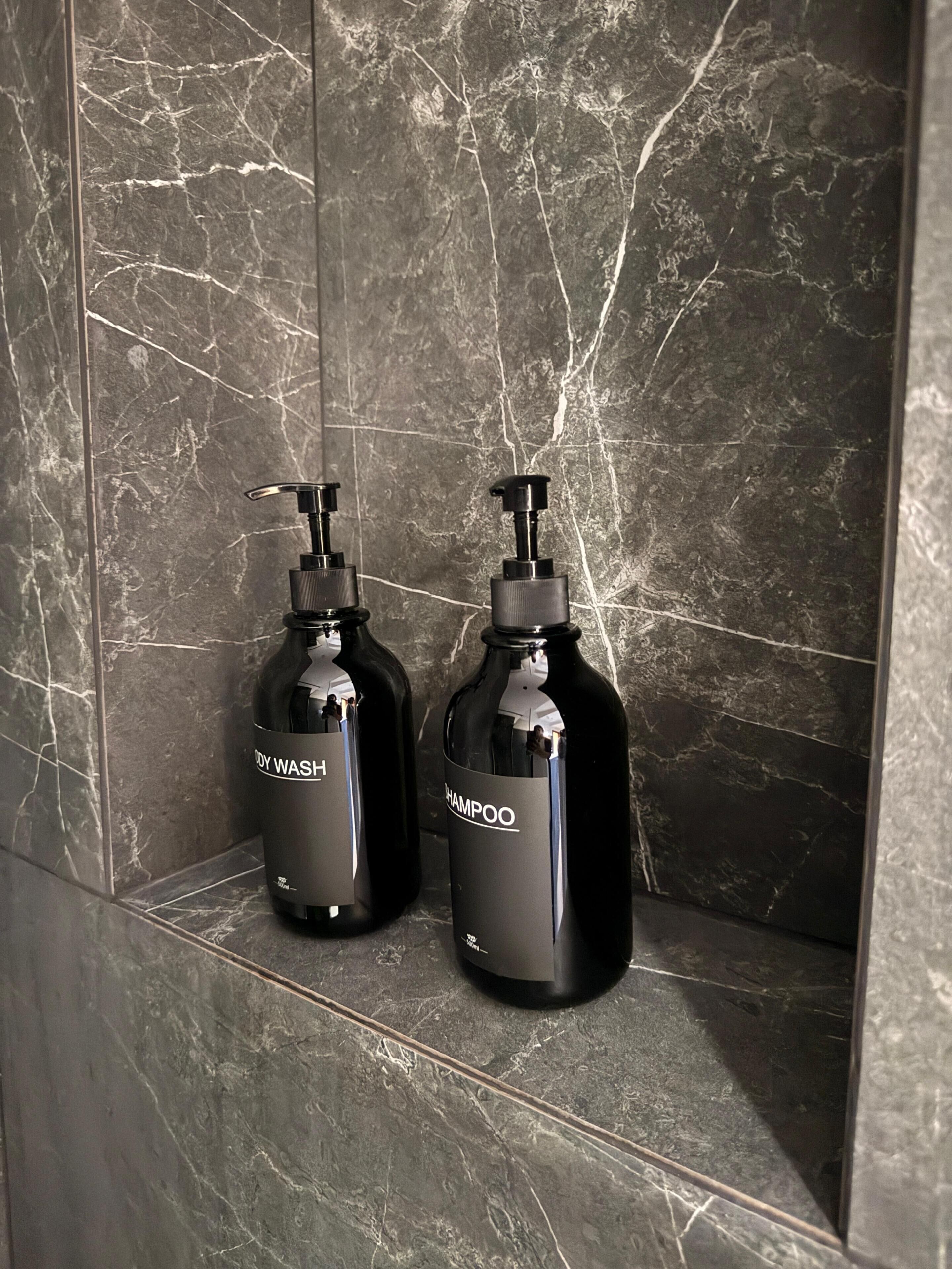 Two black bottles with pumps, labeled 'BODY WASH' and 'SHAMPOO,' on a marble-tiled wall.