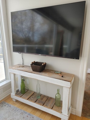Un tĂ©lĂ©viseur Ă Ă©cran plat fixĂ© Ă un mur blanc, une console en bois rustique surmontĂ©e dâun panier en osier, et deux bouteilles en verre vert.
