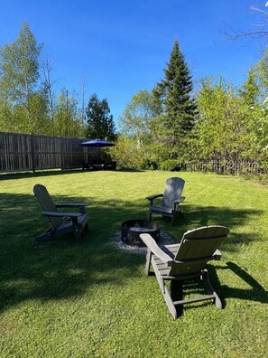 A backyard with a fire pit, two Adirondack chairs, and a wooden deck.