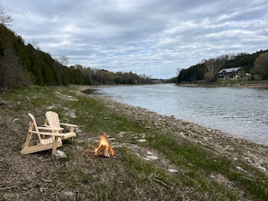 A riverside setting with a campfire and two Adirondack chairs.
