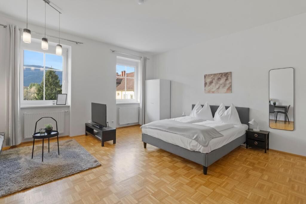 A bedroom with a bed, a chair, a TV, a radiator, and a window with a view.