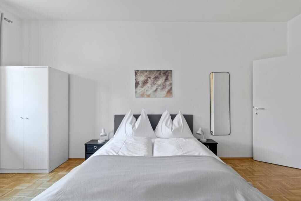 A modern hotel room with a bed, a wardrobe, a bedside table with a lamp, and a wall-mounted mirror.