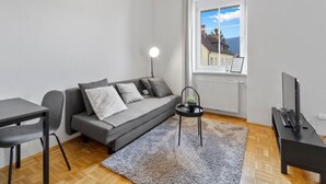 A modern living room with a sofa, a small table, a chair, a TV, and a window with a view of a house. - FarawayHomes Studio Krieglach 14 (Sommer)