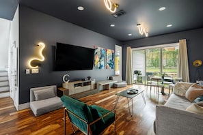 A modern living room with a flat-screen TV, artwork, a sofa, a coffee table, and a dining area with a view of greenery. - 3BR East Austin Unit A - Sleeps 10, Pet Friendly (Austin)