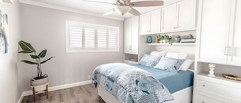 Un dormitorio con cama, ventilador de techo, armarios blancos y ventana con persianas.