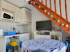 Eine kompakte Küche mit Mikrowelle, Herd und Fernseher auf einem Schrank. Es gibt einen Esstisch mit Stühlen, eine Treppe und ein kleines Regal mit Dekoration.