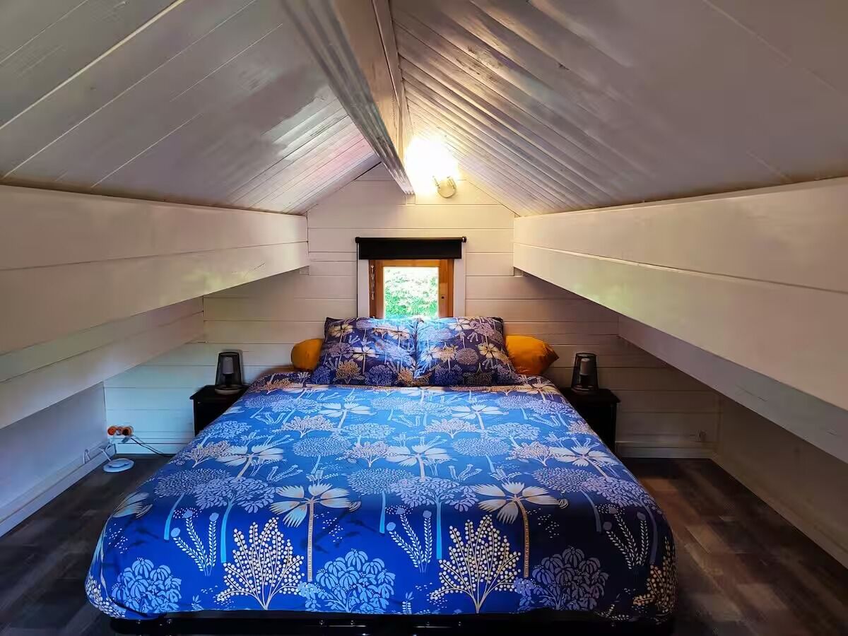 Ein Schlafzimmer mit schrägem Dach, einem Bett mit floralem Überwurf, zwei Nachttischen mit Lampen und einem Fenster mit Blick ins Grüne.