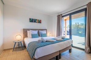 Une chambre d’hôtel avec un lit, une petite table avec une lampe, un tableau au mur et une vue à travers une porte vitrée.