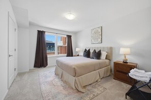 A bedroom with a large bed, a nightstand, a lamp, a window with curtains, and a door. - Luxury 2 BR townhome with EV charging (Detroit)