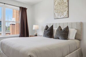 A bedroom with a large bed, a nightstand with a lamp, a window with curtains, and a decorative wall piece. - Luxury 2 BR townhome with EV charging (Detroit)