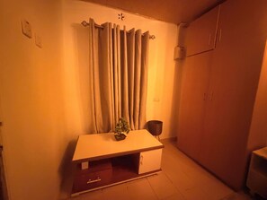 Ein Zimmer mit einem weißen Schrank, einem kleinen Tisch mit einer Pflanze, einem Vorhang und einer Wand-Klimaanlage.