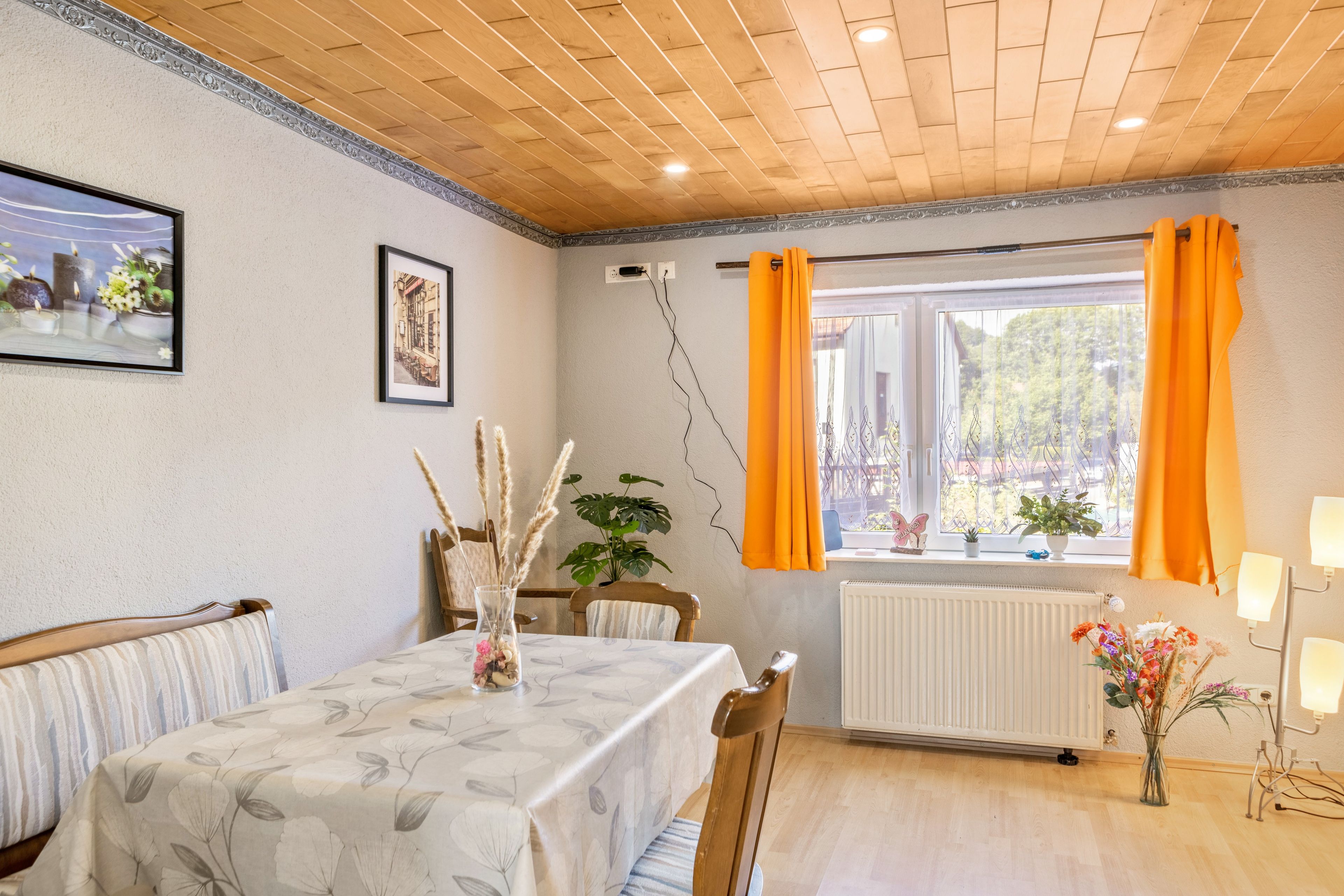 Un coin repas avec une table dressée pour deux personnes, une chaise, un radiateur et une fenêtre avec des rideaux.