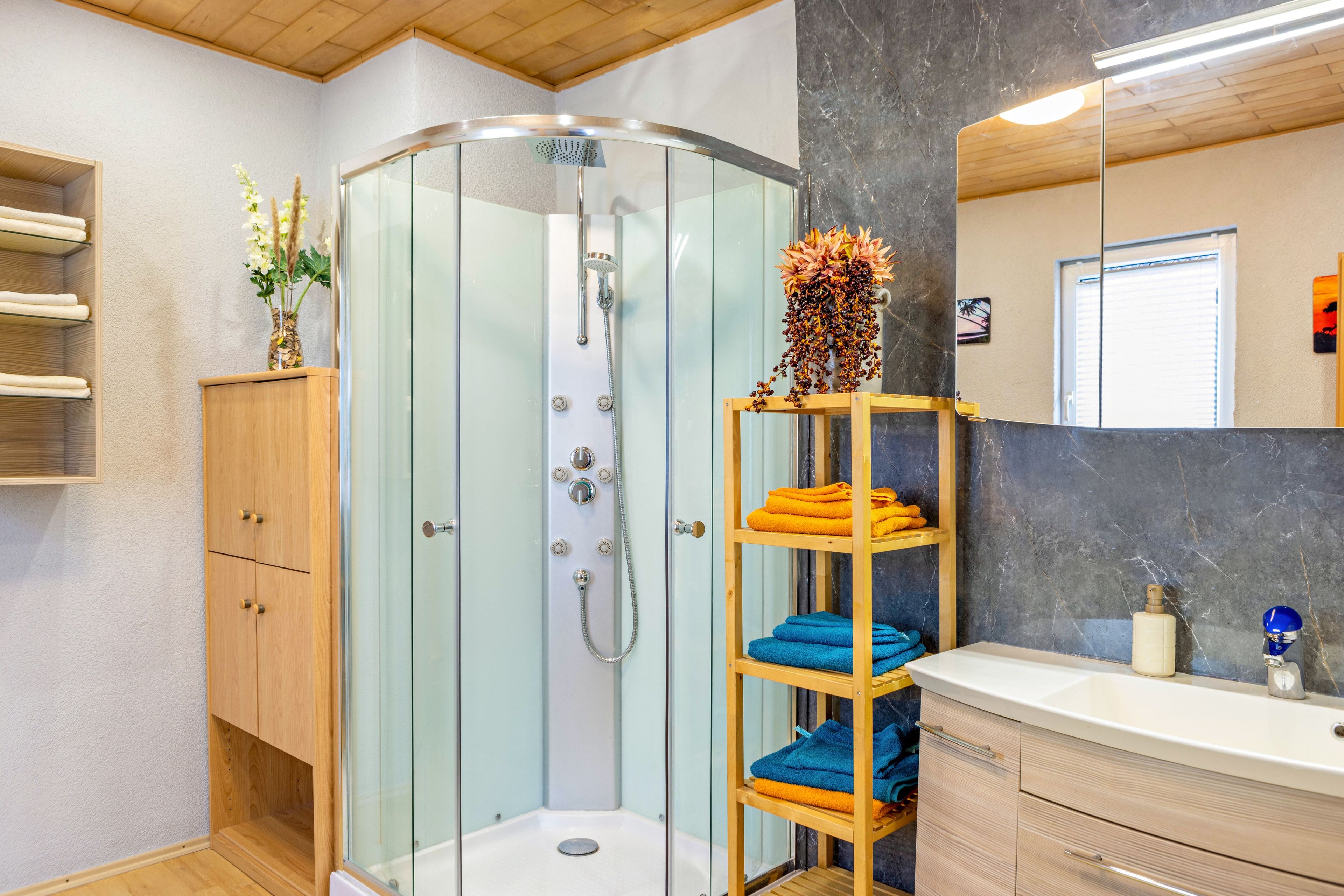 A modern bathroom with a glass-enclosed shower, wooden shelves with towels, a marble wall, and a wooden ceiling.