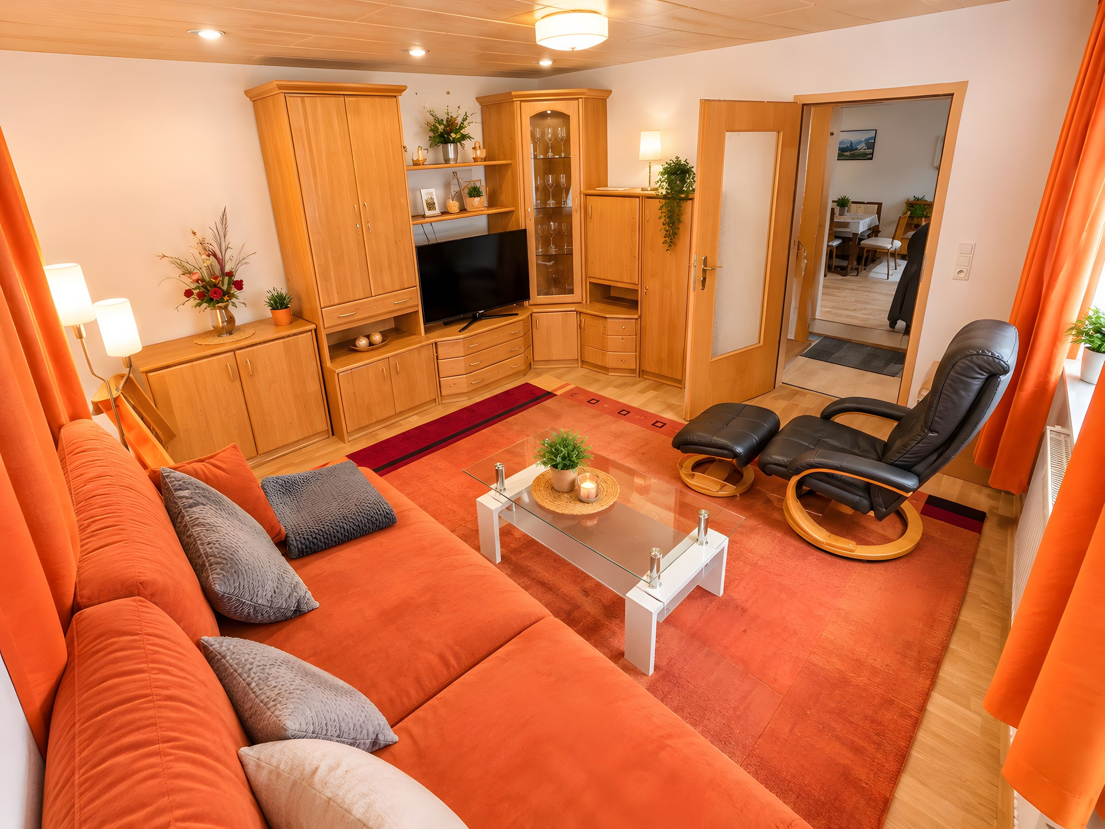 A living room with a sofa, a television, wooden cabinets, and a dining area in the background.