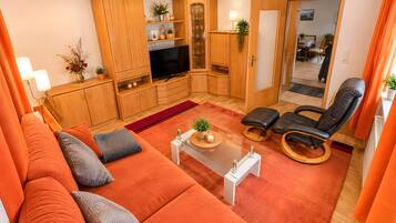 A living room with a sofa, a television, wooden cabinets, and a dining area in the background.