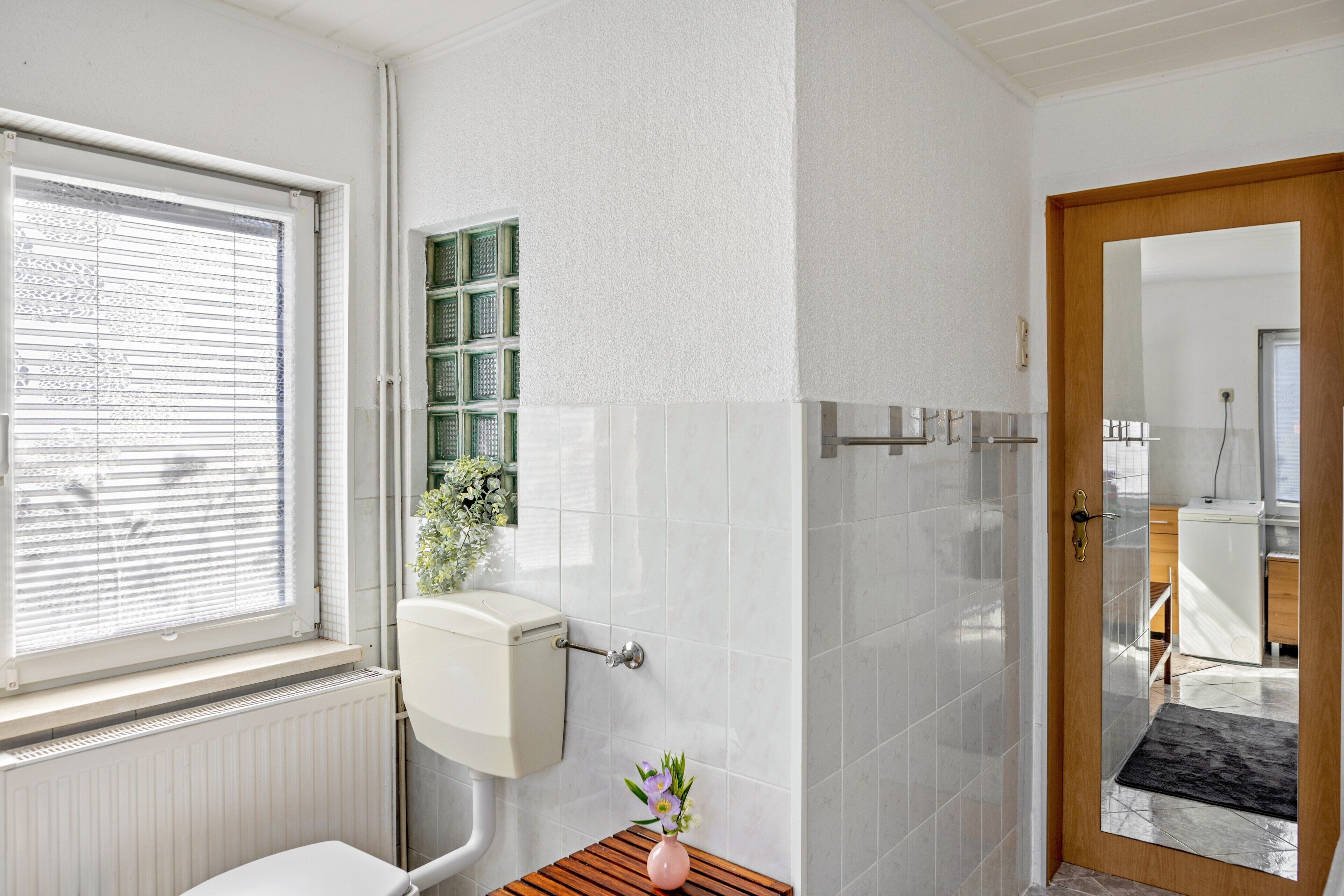 Ein Badezimmer mit Toilette, einem Fenster, einer Heizung, einer kleinen Holzablage mit einer Vase und einem Spiegel an der Tür.