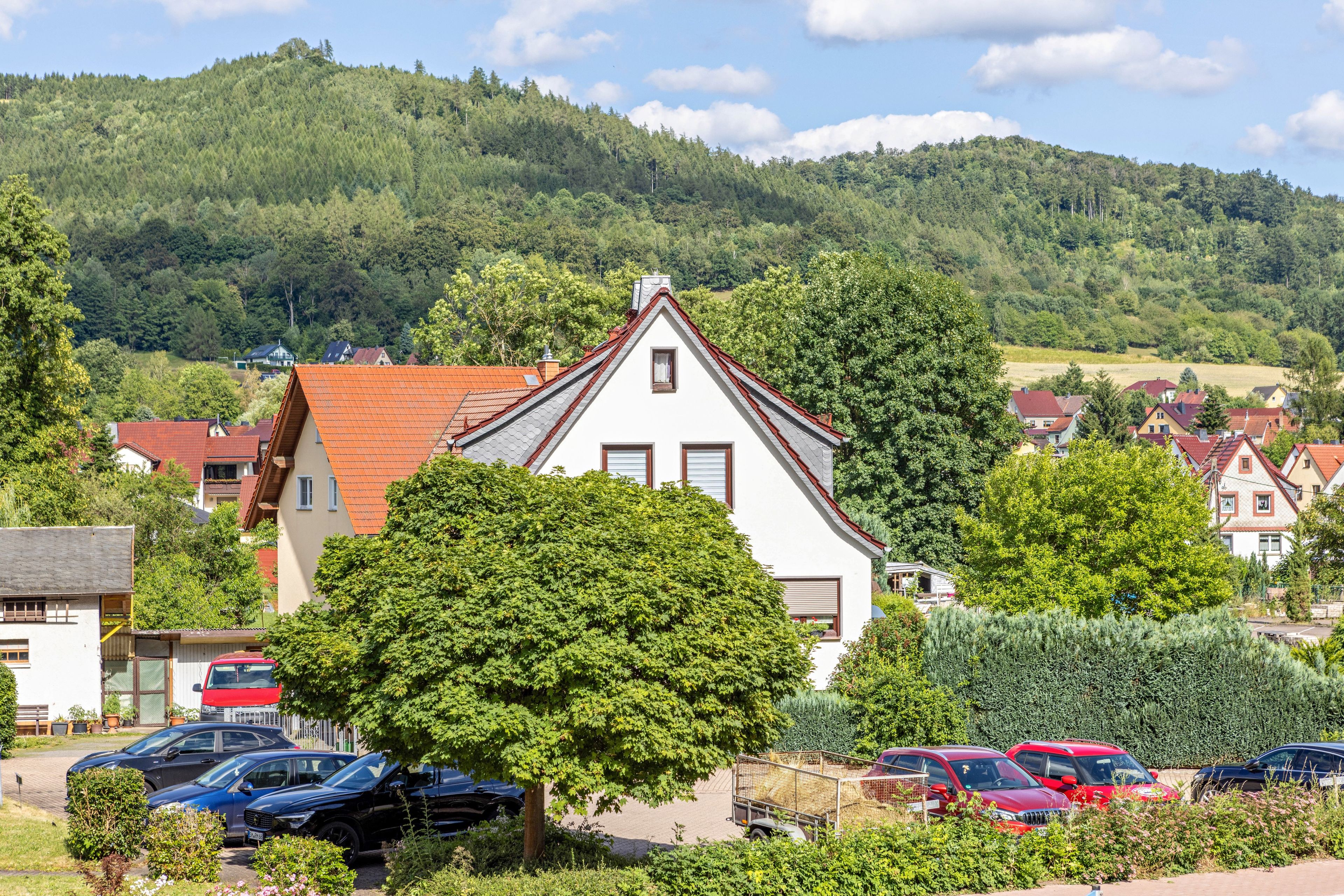 Ein weißes Gebäude mit rotem Dach, umgeben von viel Grün und parkenden Autos.