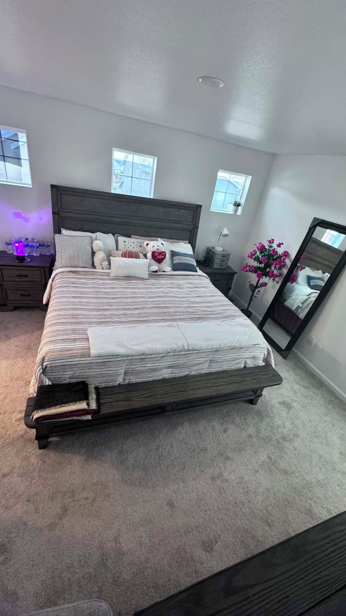 A neatly made bed in a bedroom with a headboard, a nightstand with a lamp, a dresser, and a mirror on the wall.