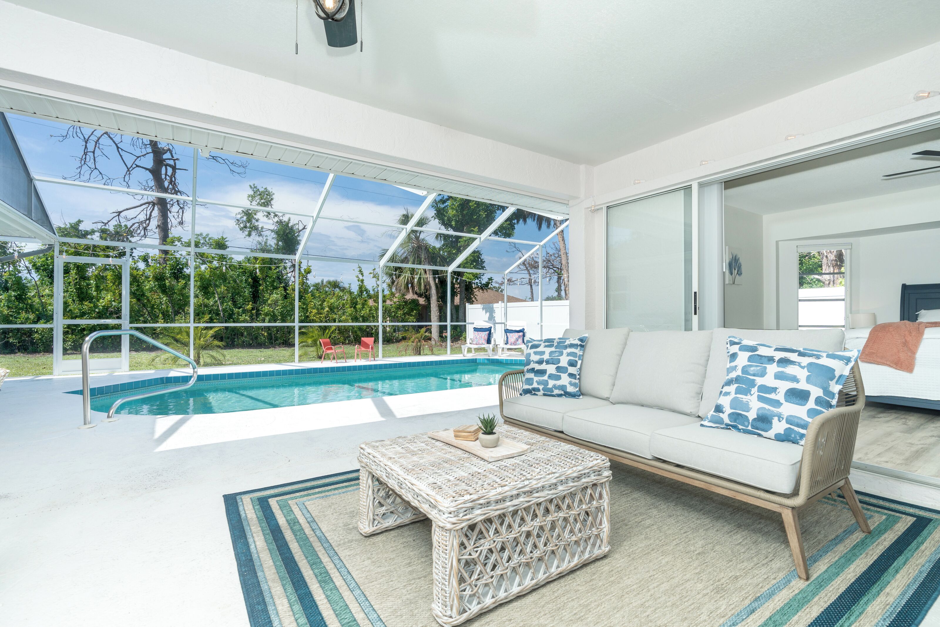 A modern living room with a large glass door leading to a pool area, a white sofa with patterned cushions, and a wicker coffee table.