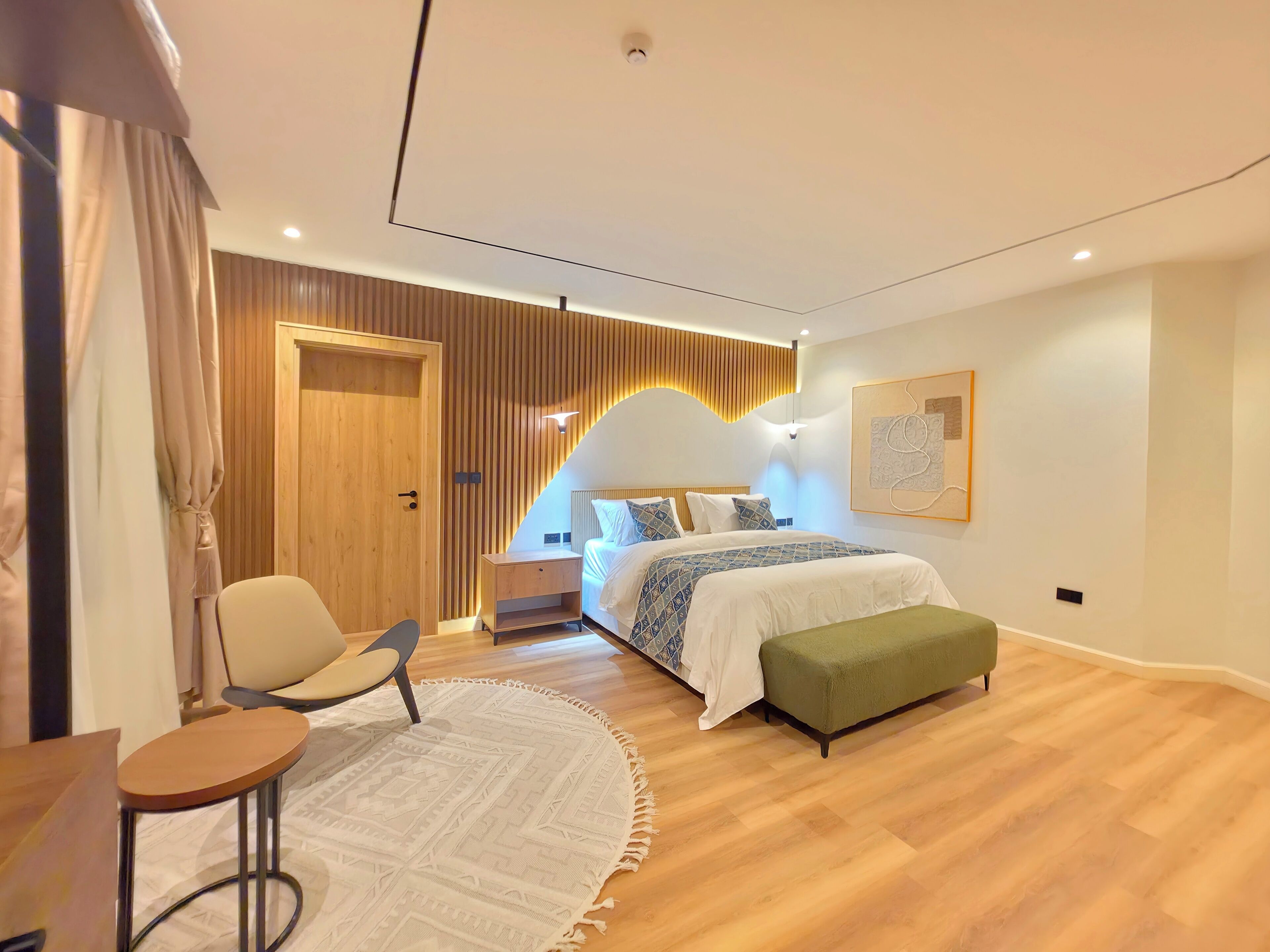 A modern bedroom with a wooden door, a bed with patterned bedding, a green bench, a round table, and a wall-mounted light fixture.