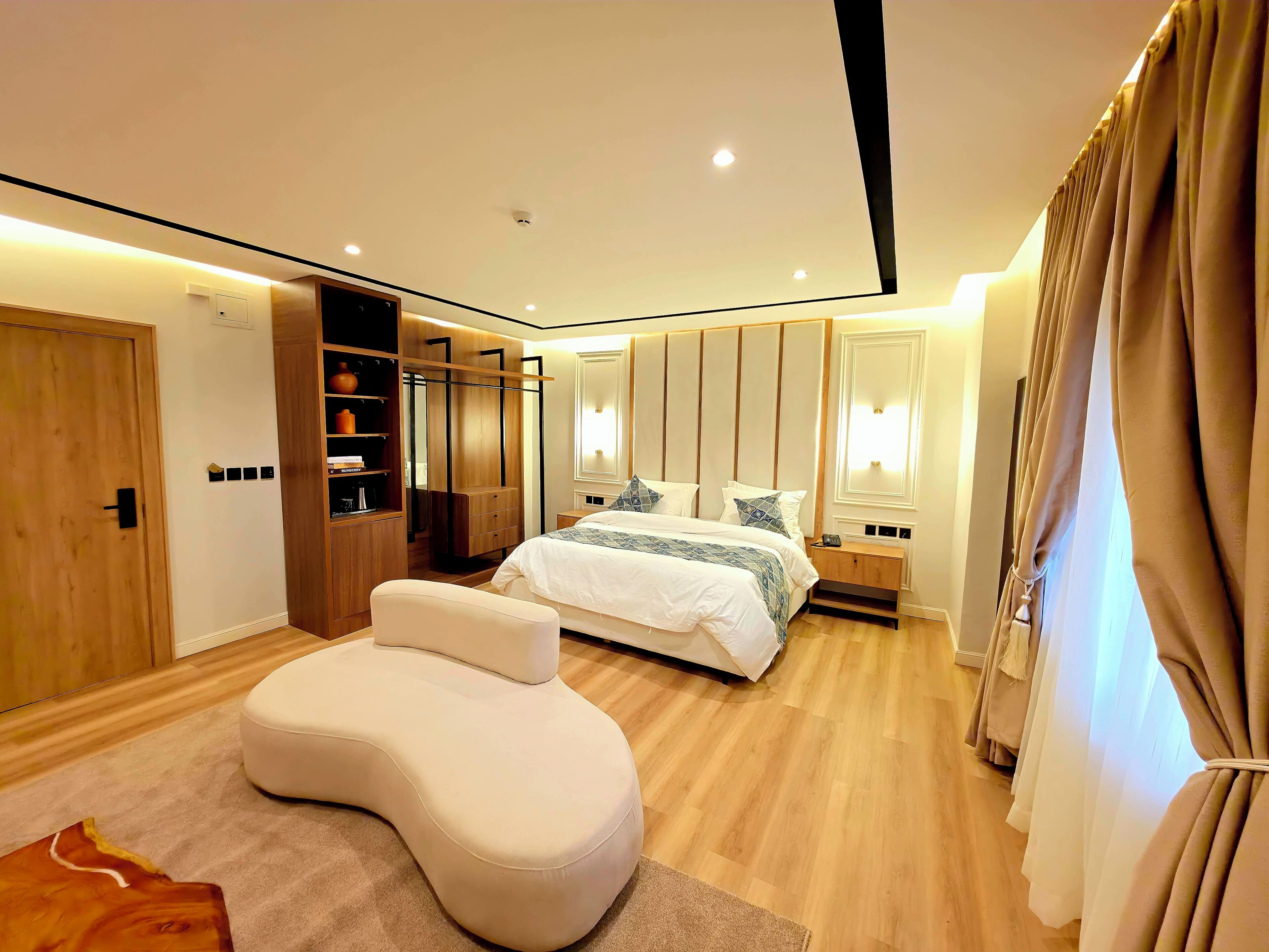 A modern bedroom with a curved white sofa, a double bed with patterned bedding, wooden wardrobe, and built-in shelves.