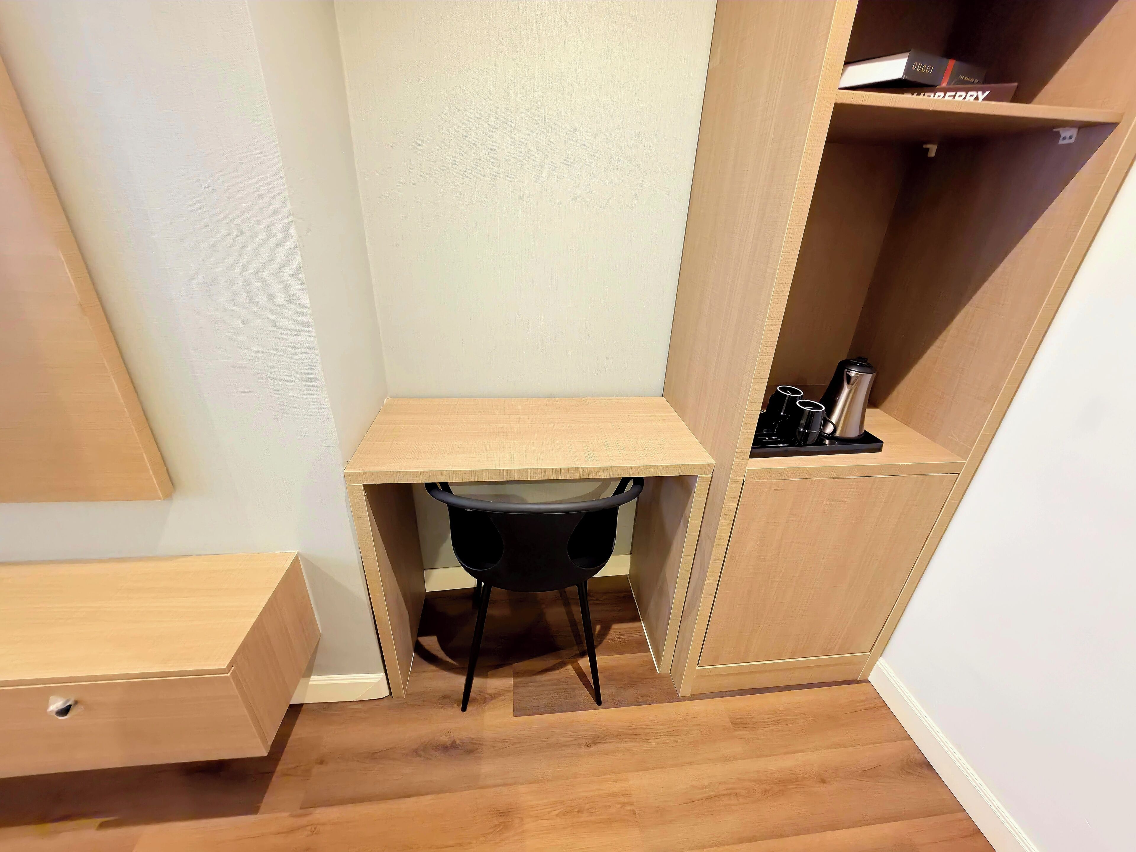 A corner room with a wooden desk, a black chair, and a shelf with books and a coffee maker.
