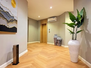A hallway with a wooden floor, a large white vase with green plants, and a painting on the wall. - Amazonia Luxury  (Abuja)