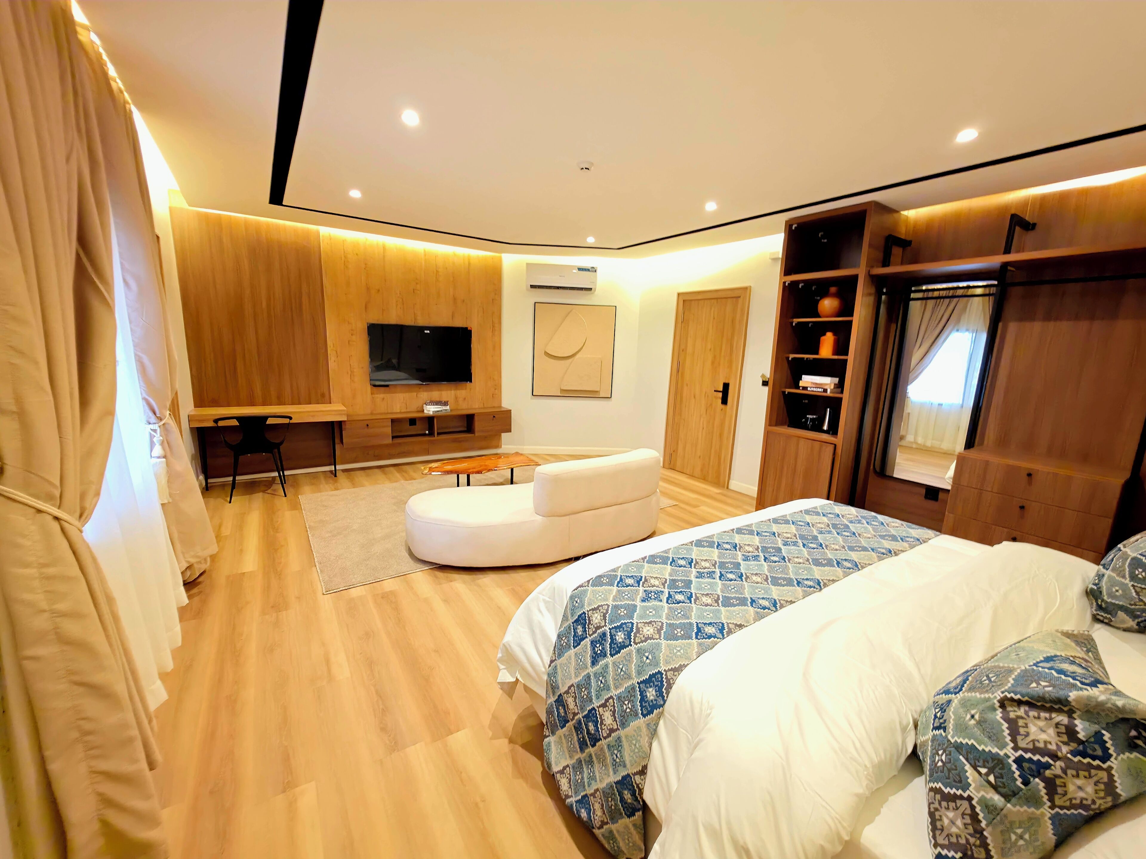 Quarto moderno com uma cama grande, televisão montada na parede, guarda-roupa de madeira e um espaço de estar com uma mesa e uma cadeira.