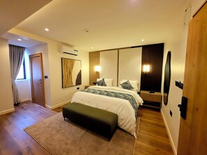 A modern bedroom with a large bed, a bench, a mirror, and a wall-mounted light fixture. - Amazonia Luxury  (Abuja)