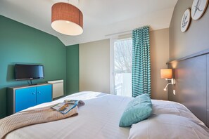 A bedroom with a bed, a television on a stand, a window with curtains, and a wall-mounted lamp. - Coastal Chalet for 6 With Pool (Courseulles-sur-Mer)