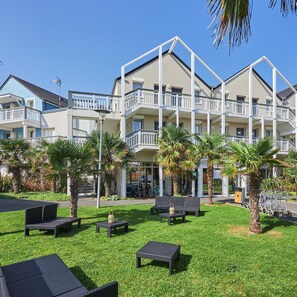 A multi-story hotel with balconies, outdoor seating, and palm trees. - Coastal Chalet for 6 With Pool (Courseulles-sur-Mer)