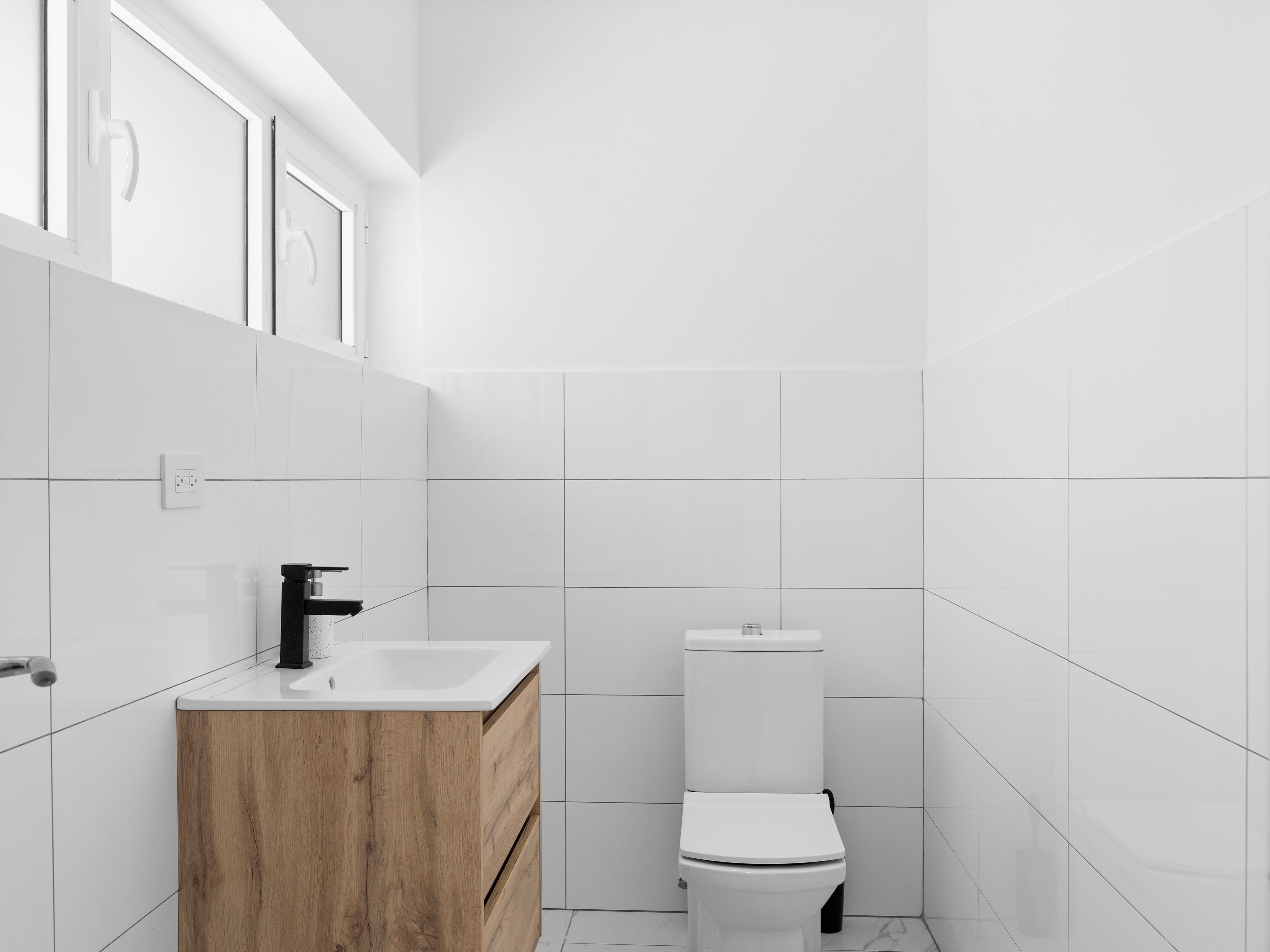 Une salle de bain moderne avec des carreaux blancs, un meuble-lavabo en bois, des toilettes blanches et un robinet noir.