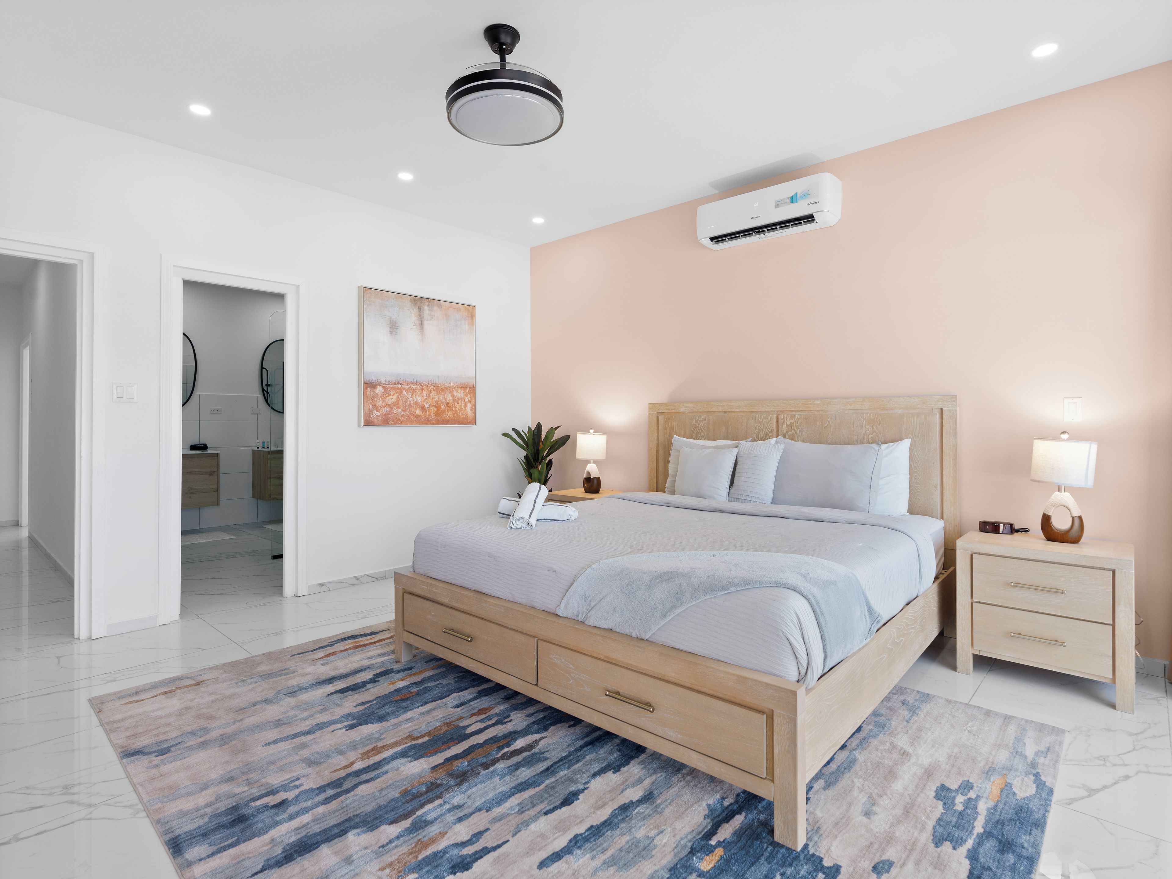 A modern bedroom with a bed, bedside tables, an air conditioning unit, and a bathroom visible through an open door.