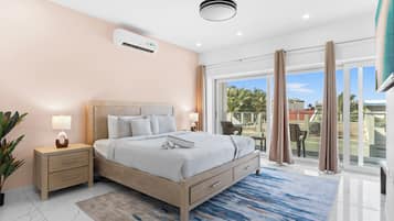 A modern bedroom with a bed, bedside tables, an air conditioner, a ceiling fan, and a view through a sliding glass door.