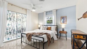 Ein Schlafzimmer mit einem Bett, einem Schreibtisch, einem Fenster mit Vorhängen, einem Deckenventilator und einem Wandbild.
