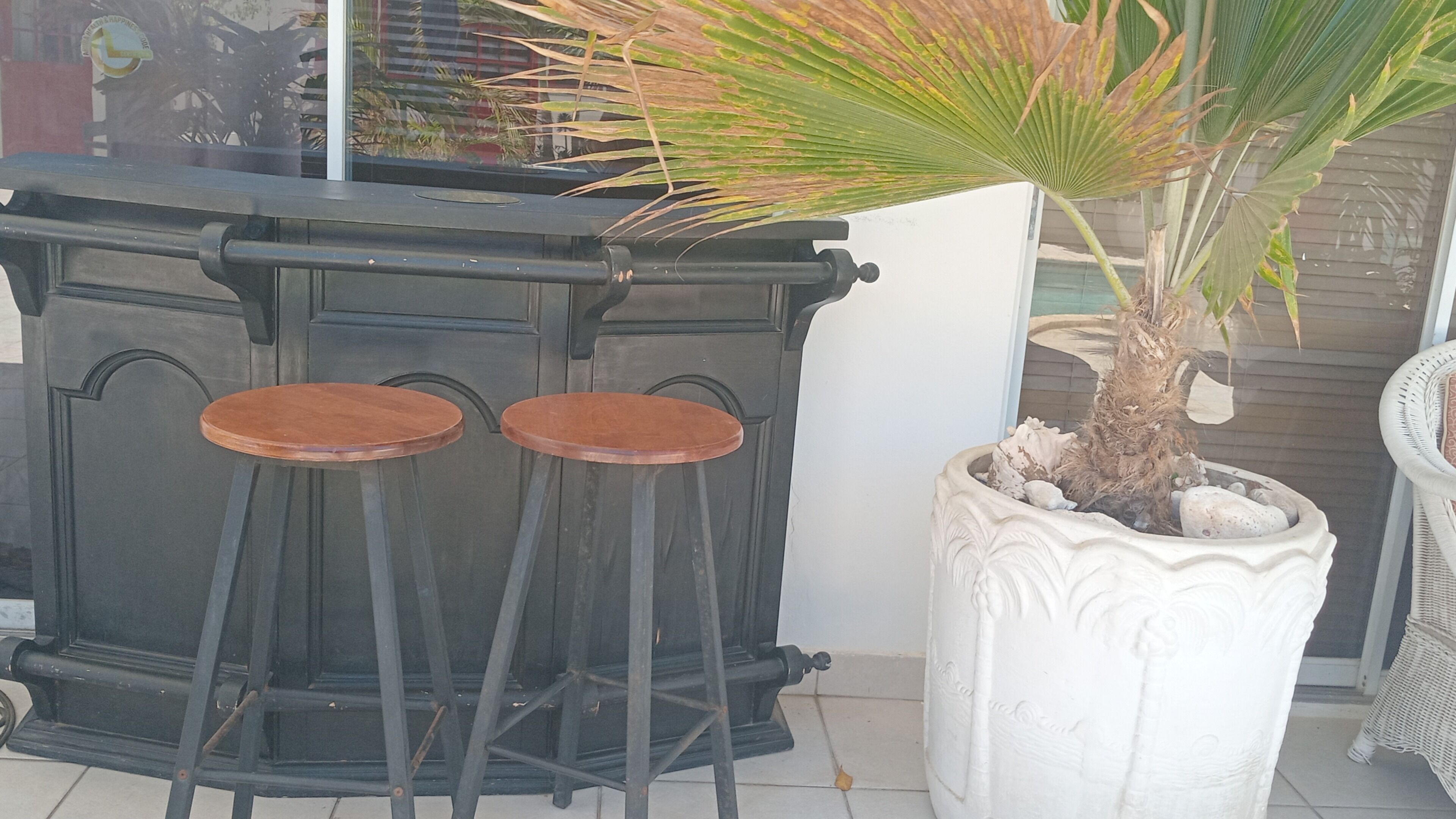 Un taburete negro frente a una pared blanca, una planta en maceta con textura y una silla de mimbre.