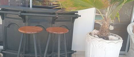 Un tabouret de bar noir est placé contre un mur blanc, à côté d’une plante en pot dont le pot a une texture particulière, et d’une chaise en osier.