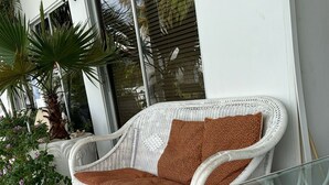 Un canapé en osier blanc agrémenté d’un coussin à motifs, une table en verre et une plante en pot, le tout placé sur une terrasse carrelée.