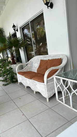 A white wicker sofa with a patterned cushion, a glass table, and a potted plant on a tiled patio. - HOSTEL WEAVING MILL ARUBA  (Oranjestad)